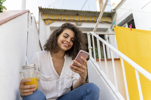 Lächelnde junge Frau sitzt mit Handy und Orangensaft auf einer Treppe - AFVF03748
