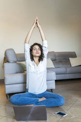 Young woman sitting on the floor at home with cell phone and laptop doing yoga - AFVF03765
