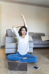 Young woman sitting on the floor at home with cell phone and laptop stretching - AFVF03767