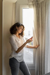 Junge Frau benutzt Handy am Fenster zu Hause - AFVF03783