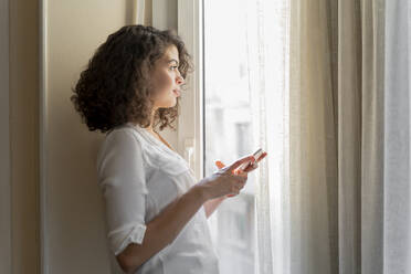Junge Frau mit Mobiltelefon, die zu Hause aus dem Fenster schaut - AFVF03785