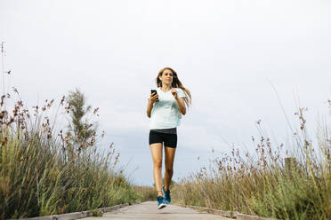 Female jogger running on wooden walkway, listening music - JRFF03645