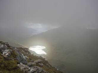 Hoher Blickwinkel auf einen See inmitten von Bergen gegen einen bewölkten Himmel bei nebligem Wetter, Schottland, UK - HUSF00057