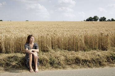 Junge Frau sitzt barfuß am Straßenrand vor einem Getreidefeld - FLLF00270