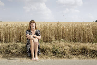Porträt einer jungen Frau, die barfuß am Straßenrand vor einem Getreidefeld sitzt - FLLF00271