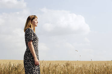 Young woman wearing dress with floral design standing in grain field - FLLF00274