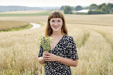 Porträt einer lächelnden jungen Frau mit einem Strauß Kamillenblüten vor einem Getreidefeld stehend - FLLF00276