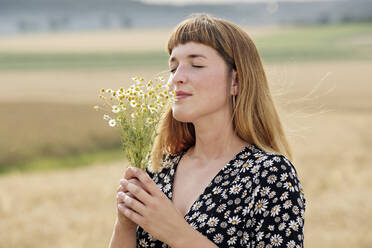 Smiling young woman with eyes closed smelling bunch of chamomile flower in nature - FLLF00279