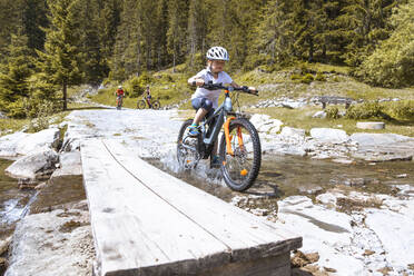 Mädchen überquert einen Gebirgsbach mit ihrem E-Mountainbike - FKF03539