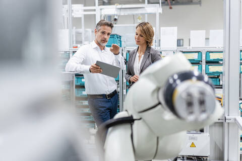 Geschäftsfrau und Mann mit Tablet im Gespräch mit Montageroboter in einer Fabrik, lizenzfreies Stockfoto