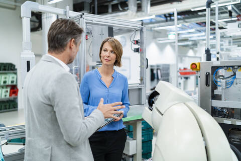 Geschäftsmann mit Tablet und Frau im Gespräch mit Montageroboter in einer Fabrik, lizenzfreies Stockfoto