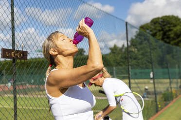 Ältere Frau im Tennisclub, die eine Pause vom Spielen macht und aus einer Wasserflasche trinkt - WPEF01725