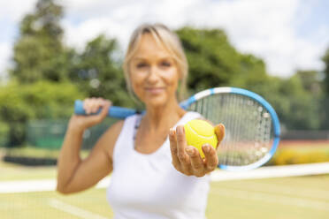 Lächelnde reife Frau hält einen Tennisball im Tennisclub - WPEF01764