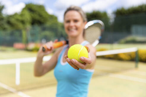 Lächelnde reife Frau hält einen Tennisball im Tennisclub - WPEF01767