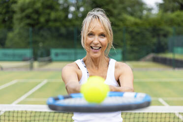 Porträt einer lachenden reifen Frau, die einen Tennisschläger und einen Ball in einem Tennisclub hält - WPEF01784