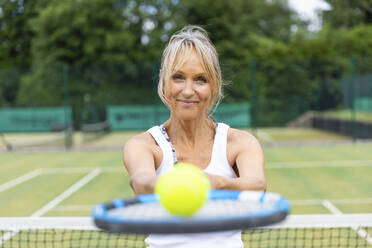 Porträt einer lächelnden reifen Frau, die einen Tennisschläger und einen Ball in einem Tennisclub hält - WPEF01786