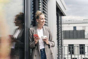 Smiling businesswoman with cell phone and coffee cup standing on roof terrace - UUF18575