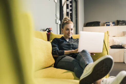 Casual businesswoman using laptop on couch in office lounge - UUF18585