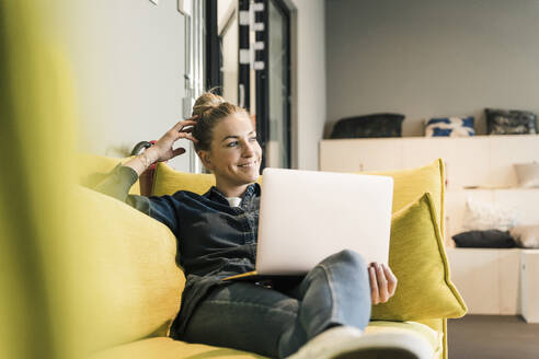 Casual businesswoman using laptop on couch in office lounge - UUF18586