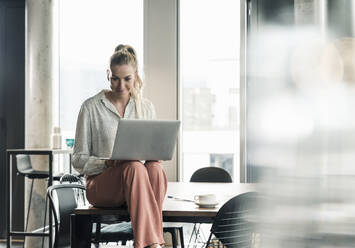 Geschäftsfrau sitzt auf einem Tisch im Büro und benutzt einen Laptop - UUF18600