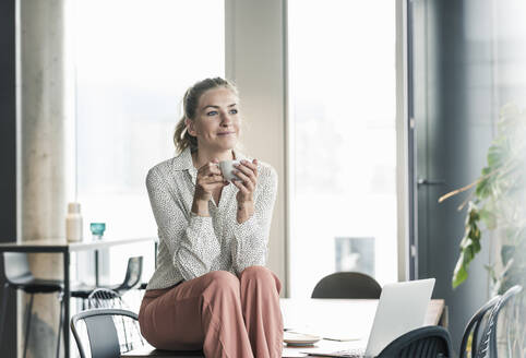 Lächelnde Geschäftsfrau sitzt auf einem Tisch im Büro und macht eine Kaffeepause - UUF18604