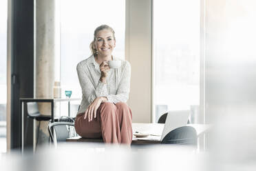 Smiling businesswoman sitting on table in office having a coffee break - UUF18605