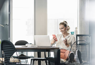 Smiling businesswoman sitting at table in office using cell phone - UUF18610