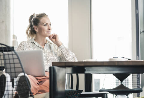 Lächelnde Geschäftsfrau sitzt am Tisch im Büro mit Laptop - UUF18612