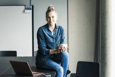 Portrait of businesswoman sitting on table in office having a coffee break - UUF18616