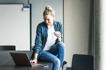 Geschäftsfrau mit einer Tasse Kaffee auf einem Tisch im Büro sitzend und einen Laptop benutzend - UUF18617