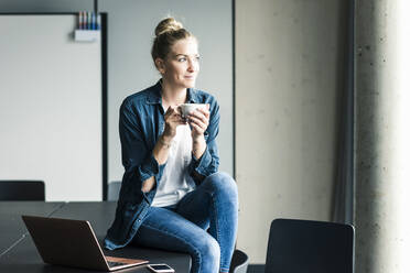 Lächelnde Geschäftsfrau sitzt auf einem Tisch im Büro und macht eine Kaffeepause - UUF18618