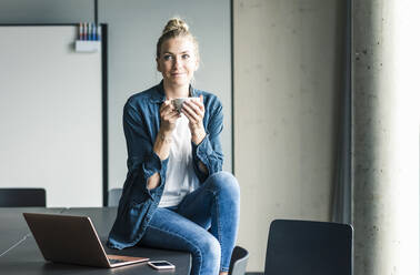 Porträt einer Geschäftsfrau, die auf einem Tisch im Büro sitzt und eine Kaffeepause macht - UUF18619