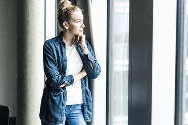 Businesswoman standing at the window in office looking out - UUF18620