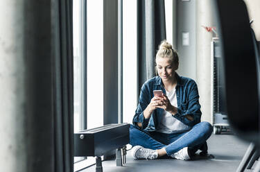 Geschäftsfrau sitzt auf dem Boden im Büro und benutzt ein Mobiltelefon - UUF18621