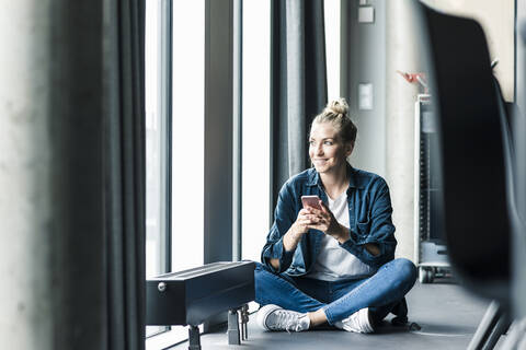 Lächelnde Geschäftsfrau auf dem Boden sitzend im Büro mit Mobiltelefon, lizenzfreies Stockfoto