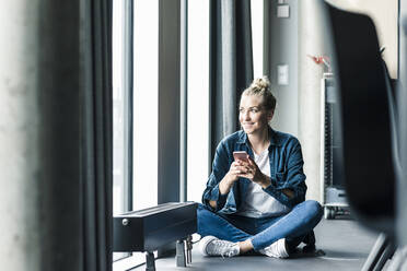 Lächelnde Geschäftsfrau auf dem Boden sitzend im Büro mit Mobiltelefon - UUF18622