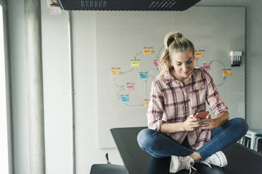 Casual businesswoman sitting on table in office using cell phone - UUF18634