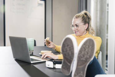 Lächelnde, lässige Geschäftsfrau, die an einem Tisch im Büro sitzt, einen Laptop benutzt und ein Sandwich isst - UUF18638