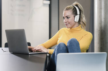 Casual businesswoman sitting at table in office using laptop and wearing headphones - UUF18643