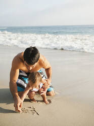 Mixed race father and daughter writing in sand together - BLEF14263