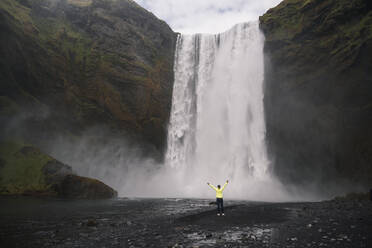 Junge Frau steht mit erhobenen Armen vor den Skogafoss-Wasserfällen, Island - UUF18660