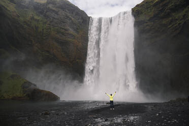 Young woman standing in front of Skogafoss waterfalls with raised arms, Iceland - UUF18660