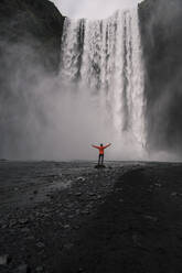 Mature man standing in front of Skogafoss waterfalls with raised arms, Iceland - UUF18664