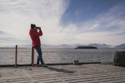 Junge Frau, die auf einem Steg steht und durch ein Fernglas schaut, Südost-Island - UUF18728
