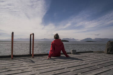 Junge Frau sitzt auf einem Steg und blickt auf das Meer, Südostisland - UUF18729