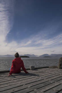 Junge Frau sitzt auf einem Steg und blickt auf das Meer, Südostisland - UUF18730