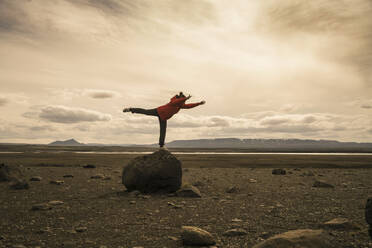 Junge Frau balanciert auf einem Bein auf einem Felsen im vulkanischen Hochland von Island - UUF18779