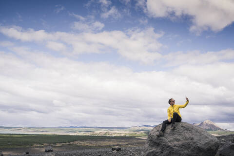 Junge Frau sitzt auf einem Felsen am Myvatn, Island, und macht Selfies, lizenzfreies Stockfoto