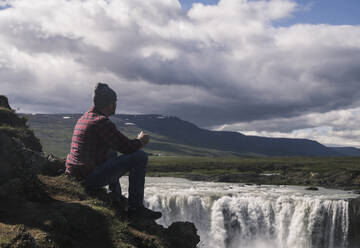 Mann sitzt mit einer Tasse Kaffee und schaut auf die Godafoss-Wasserfälle, Island - UUF18793