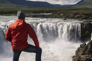 Mann betrachtet die Godafoss-Wasserfälle, Island, mit einem Fernglas - UUF18794